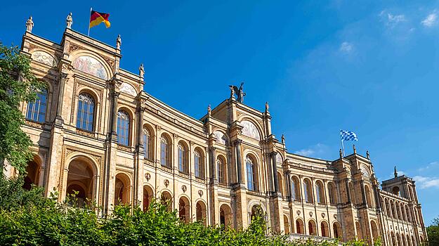 Großer Raum mit leeren, rot gepolsterten Sitzreihen und hellen Wänden. Hinter einem Rednerpult sind Flaggen und ein großes Wappen an der Wand zu sehen. 